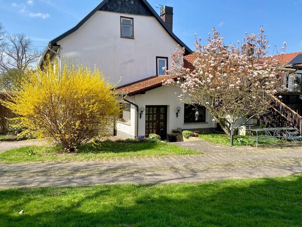 Ein zweigeschossiges Haus mit weißer Fassade, einem roten Ziegeldach und einer kleinen Veranda mit einer Bank.