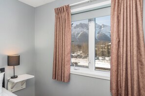 Une chambre avec un lit, une lampe de chevet, une fenêtre avec des rideaux et une vue sur des montagnes enneigées.