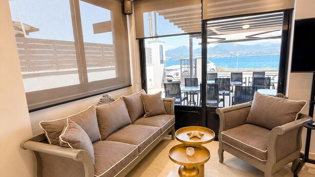 Una sala de estar moderna con sofá, sillones y una mesa de centro, ventanas amplias con vista al mar, y un balcón con mobiliario exterior.