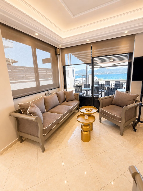 A modern living room with a sofa, armchairs, and a coffee table, large windows with a view of the sea, and a balcony with outdoor seating.