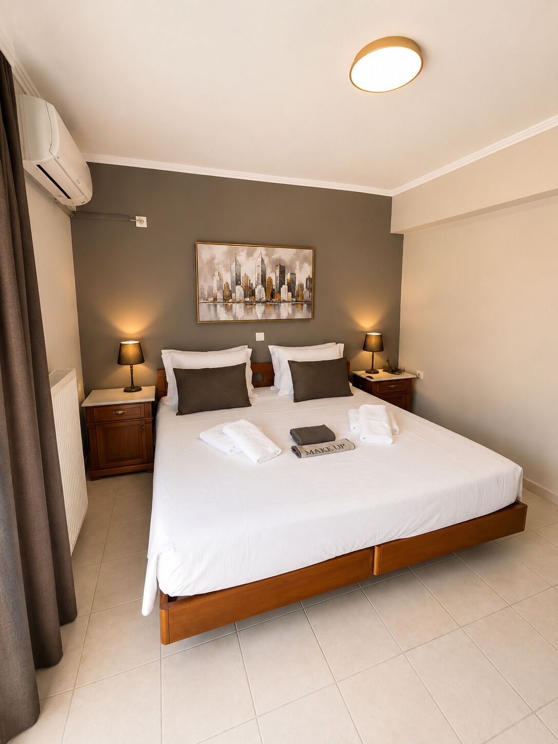 Quarto de hotel com uma cama grande, duas mesas de cabeceira, ar condicionado fixado na parede e uma pintura com vista para a cidade.