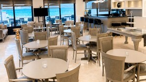 Un comedor moderno con mesas y sillas redondas, una zona de bar y grandes ventanales con vista al mar.