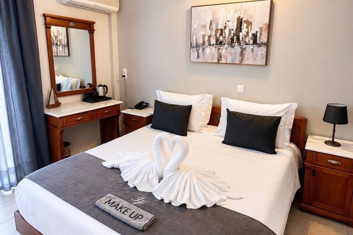 A hotel room with a large bed, a bedside table, a desk, a chair, a mirror, a painting, and a wall-mounted light fixture.