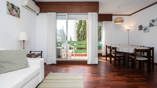 Une pièce avec un plafond en bois, un canapé blanc, une table à manger avec des chaises, un banc vert à l’extérieur et une vue sur les arbres par la fenêtre.