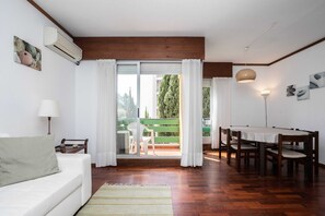Una habitación con techo de madera, un sofá blanco, una mesa de comedor con sillas, un banco verde afuera y vistas a árboles por la ventana.