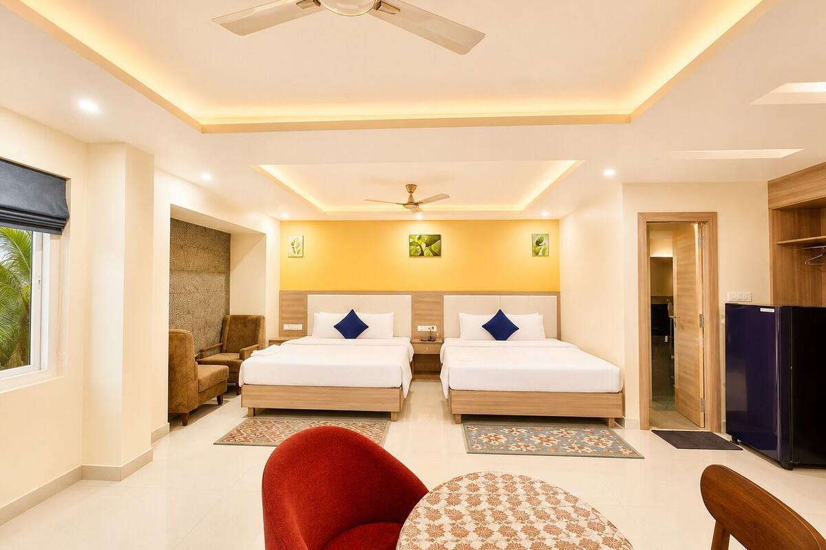 A hotel room with two beds, a ceiling fan, a refrigerator, and a dining area with a table and chairs.