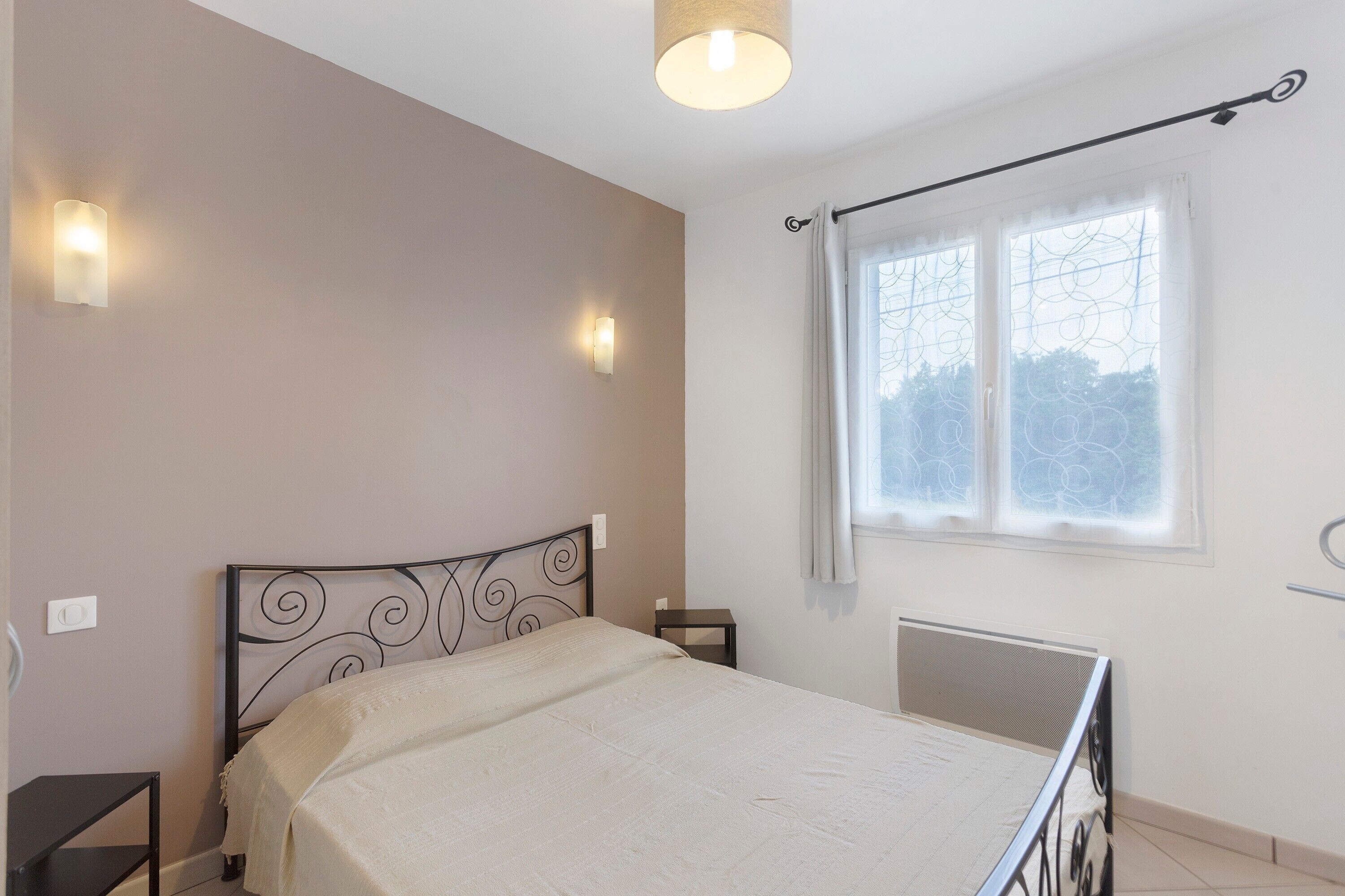 Un dormitorio con cama, mesitas de noche, una ventana con cortinas e iluminación en la pared.