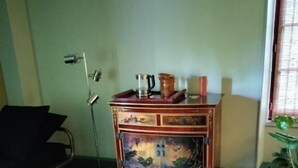 A room with a green ottoman, a wooden side table, a vintage cabinet, a floor lamp, and a window with blinds. - Piece of Pai (Sainte-Hermine)
