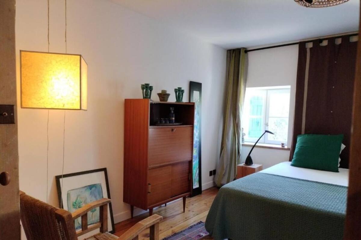 A bedroom with a bed, a wooden dresser, a chair, a lamp, and a window with curtains.