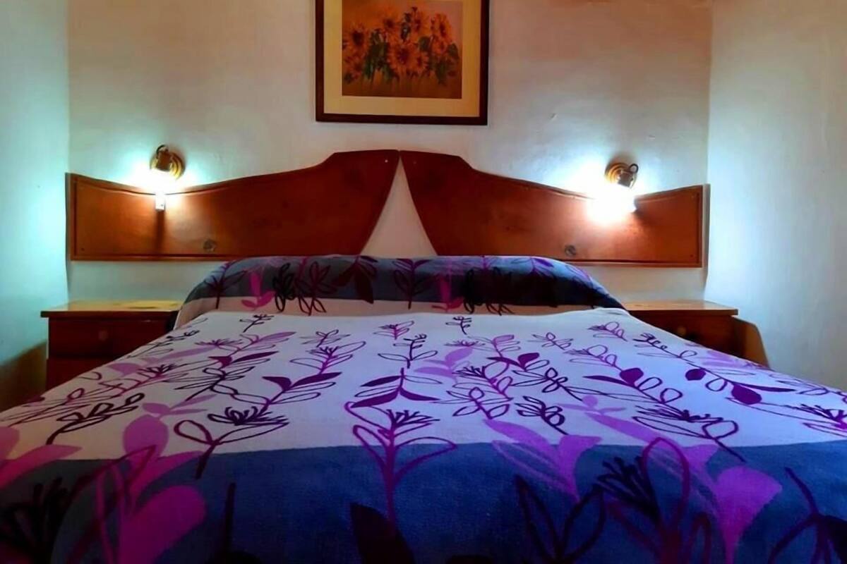 Una cama con una colcha de estampado floral, un cabecero de madera, dos lámparas de noche y un cuadro enmarcado en la pared.