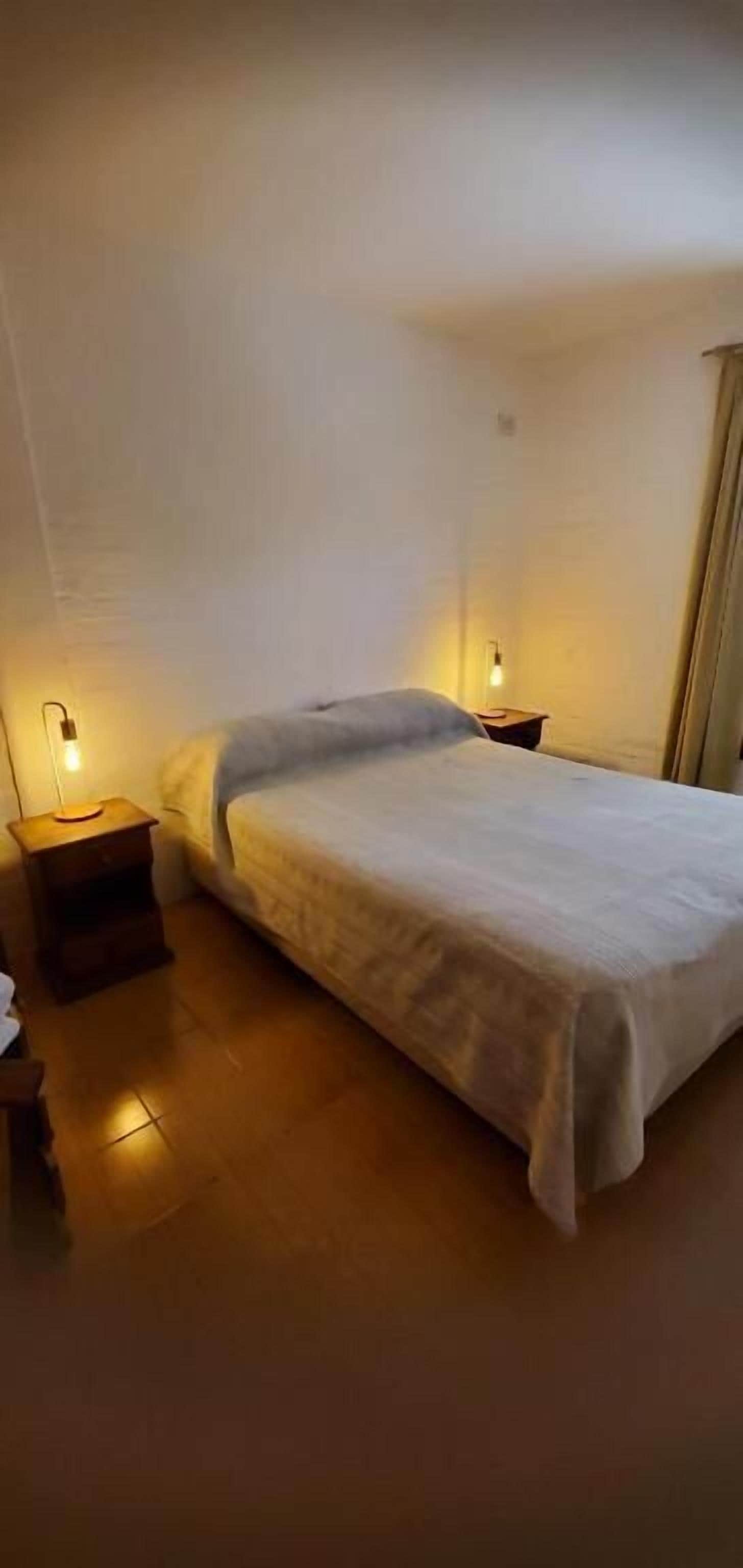 Une petite chambre à coucher peu éclairée, avec un lit simple, une table de chevet et une lampe fixée au mur.