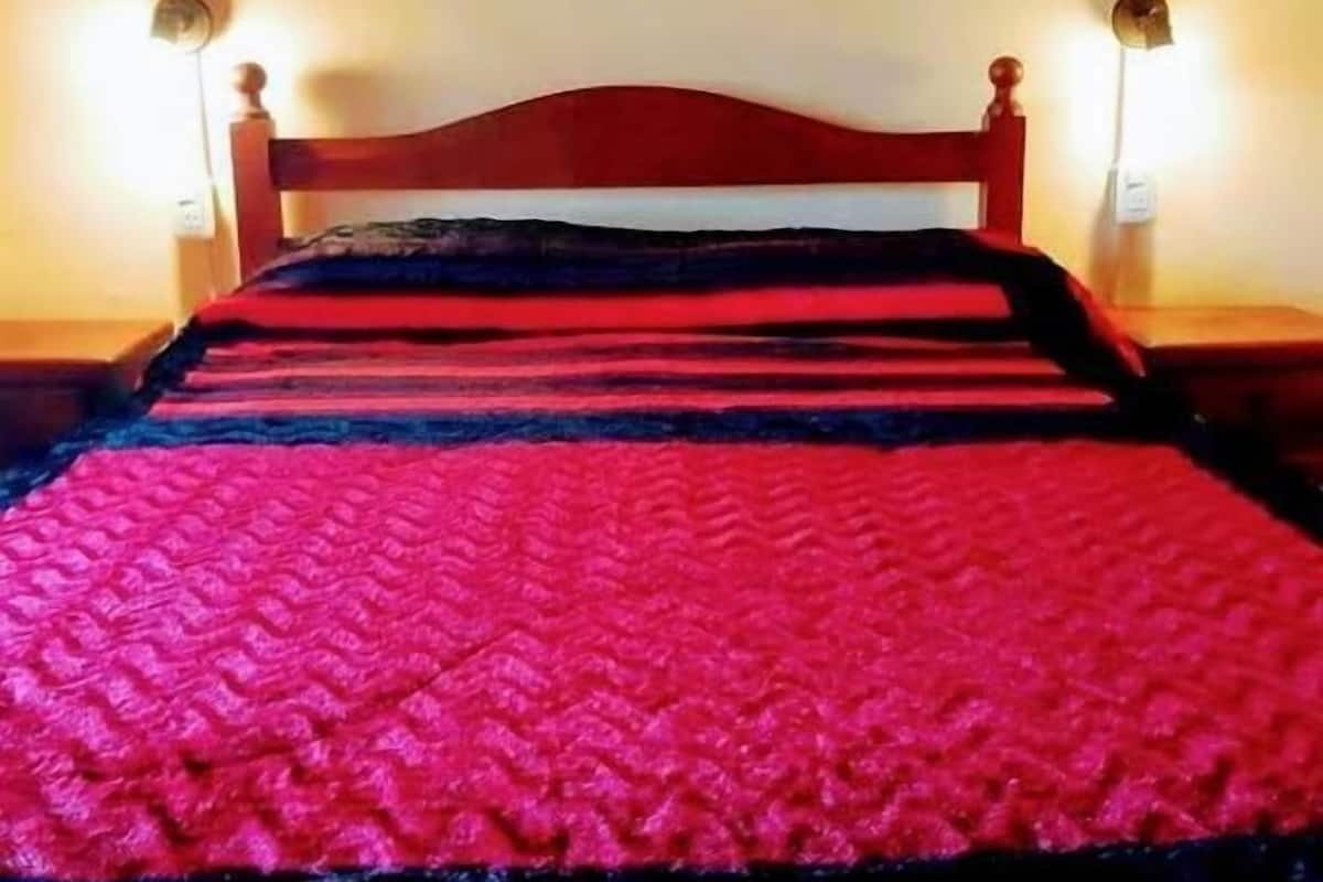 Una cama con una colcha roja y negra, dos lámparas de pared y una pared con tres cuadros de flores.