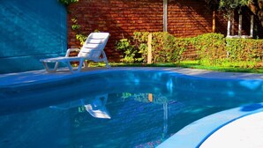 Una piscina con una tumbona blanca, un muro de ladrillo y follaje al fondo.
