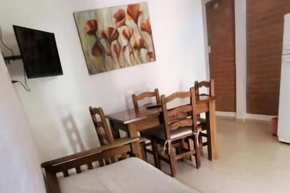 Una habitación con cama, una mesa de comedor con sillas, un televisor montado en la pared y un cuadro.