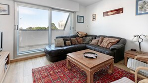 Ein Wohnzimmer mit einem schwarzen Ledersofa, einem Holztisch, einem roten Teppich mit Muster und einer Glasschiebetür, die zu einem Außenbalkon führt.