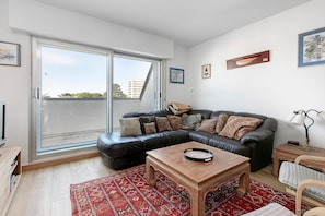 Een woonkamer met een zwarte leren bank, een houten salontafel, een rood geweven vloerkleed en een schuifdeur die leidt naar een buitenbalkon.