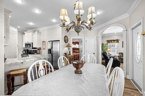 A dining area with a large table, white chairs, a chandelier, and a kitchen in the background. - Short Walk to Beach, Waterpark & Golf Cart (Myrtle Beach)