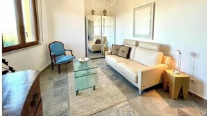 Een woonkamer met een glazen salontafel, een blauwe fauteuil, een witte bank, een kroonluchter en een groot raam.
