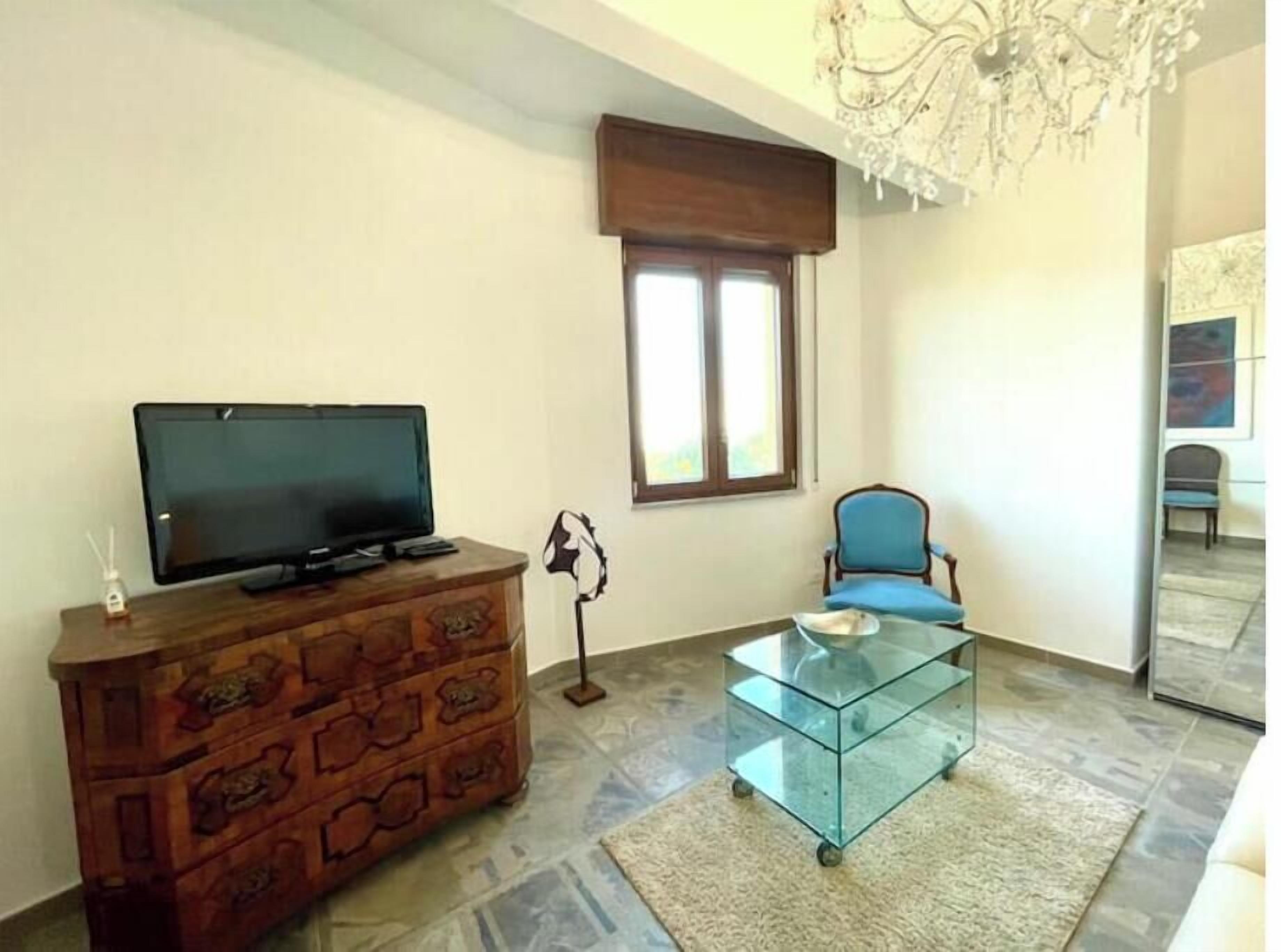 Una sala de estar con un televisor sobre un mueble de madera, una mesa de centro de vidrio, una silla azul, una ventana con persianas y una araña.