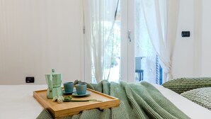 Une chambre à coucher avec un lit, un plateau en bois sur lequel se trouvent une machine à café et des tasses, une couverture verte et des rideaux en voile.
