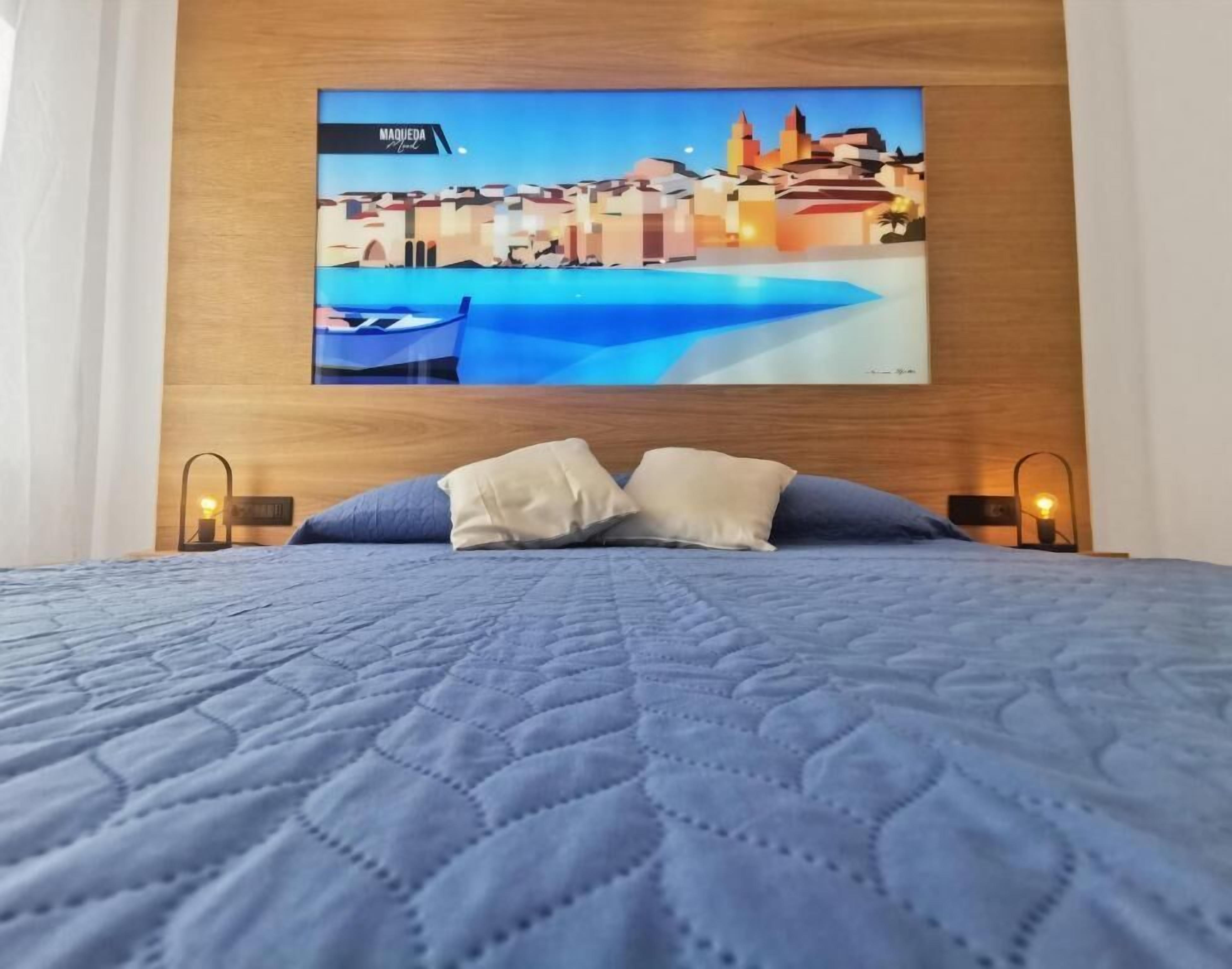 Ein Bett mit einer gesteppten blauen Decke, zwei Kissen und einem an der Wand montierten Fernseher, der eine Stadtansicht zeigt.