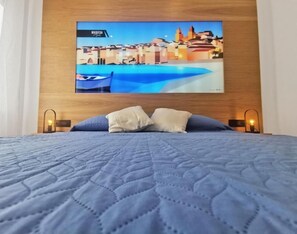 A bed with a quilted blue blanket, two pillows, and a wall-mounted TV displaying a cityscape. - Maqueda Mood (Palermo)