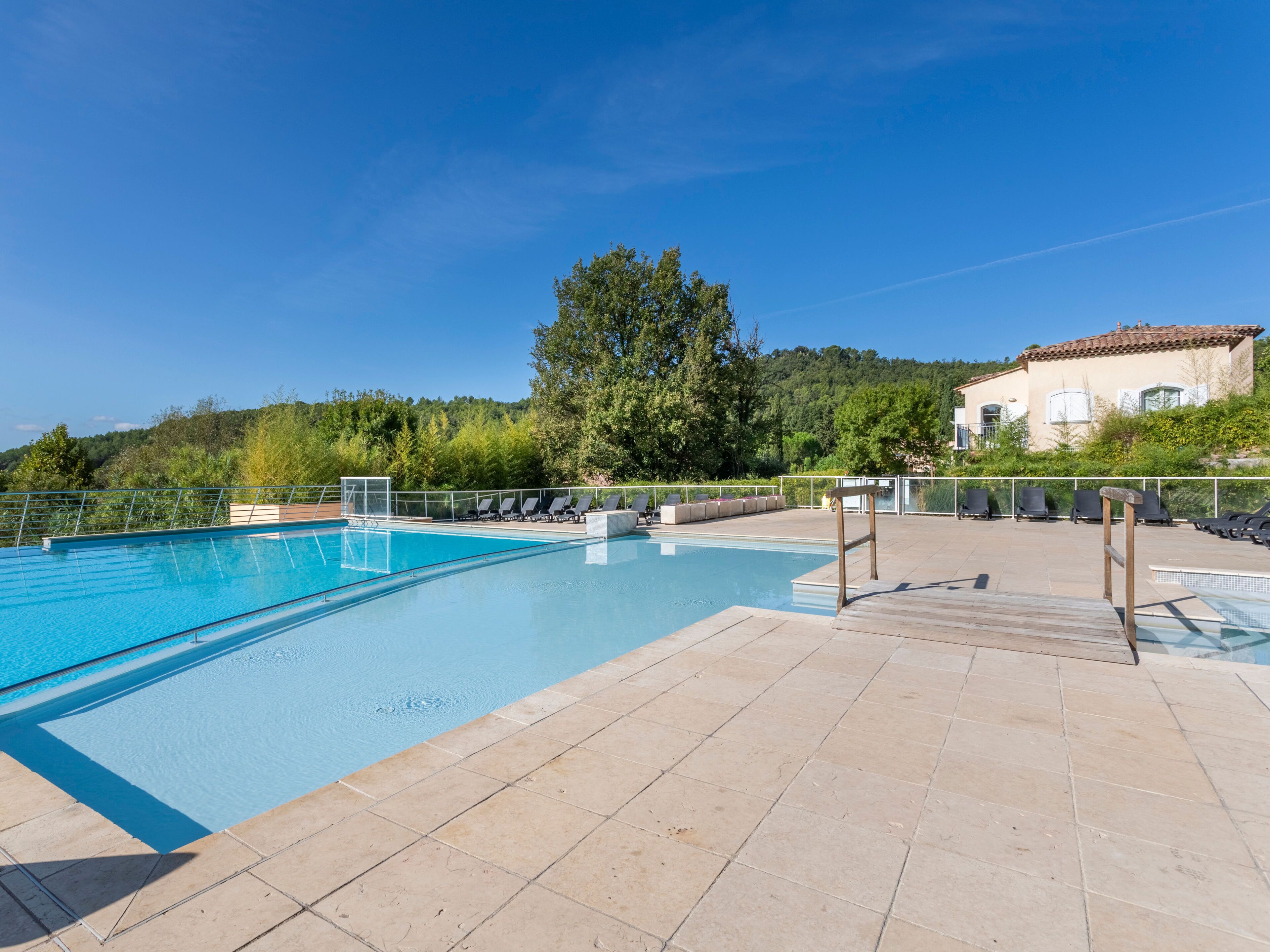 A swimming pool with a tiled deck, lounge chairs, and a view of a hillside with trees and a building.