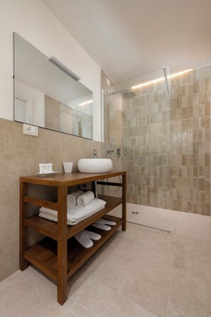A modern bathroom with a wooden vanity, a large mirror, a shower with a glass door, and beige tiles. - Terramarina Boutique Hotel (SIROLO)