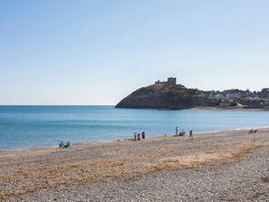 A pebbly beach with a castle on a hill, clear blue water, and a few people. - 61A High Street (Criccieth)