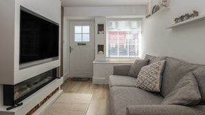A living room with a flat-screen TV, a grey sofa, a white door, and a window with blinds.