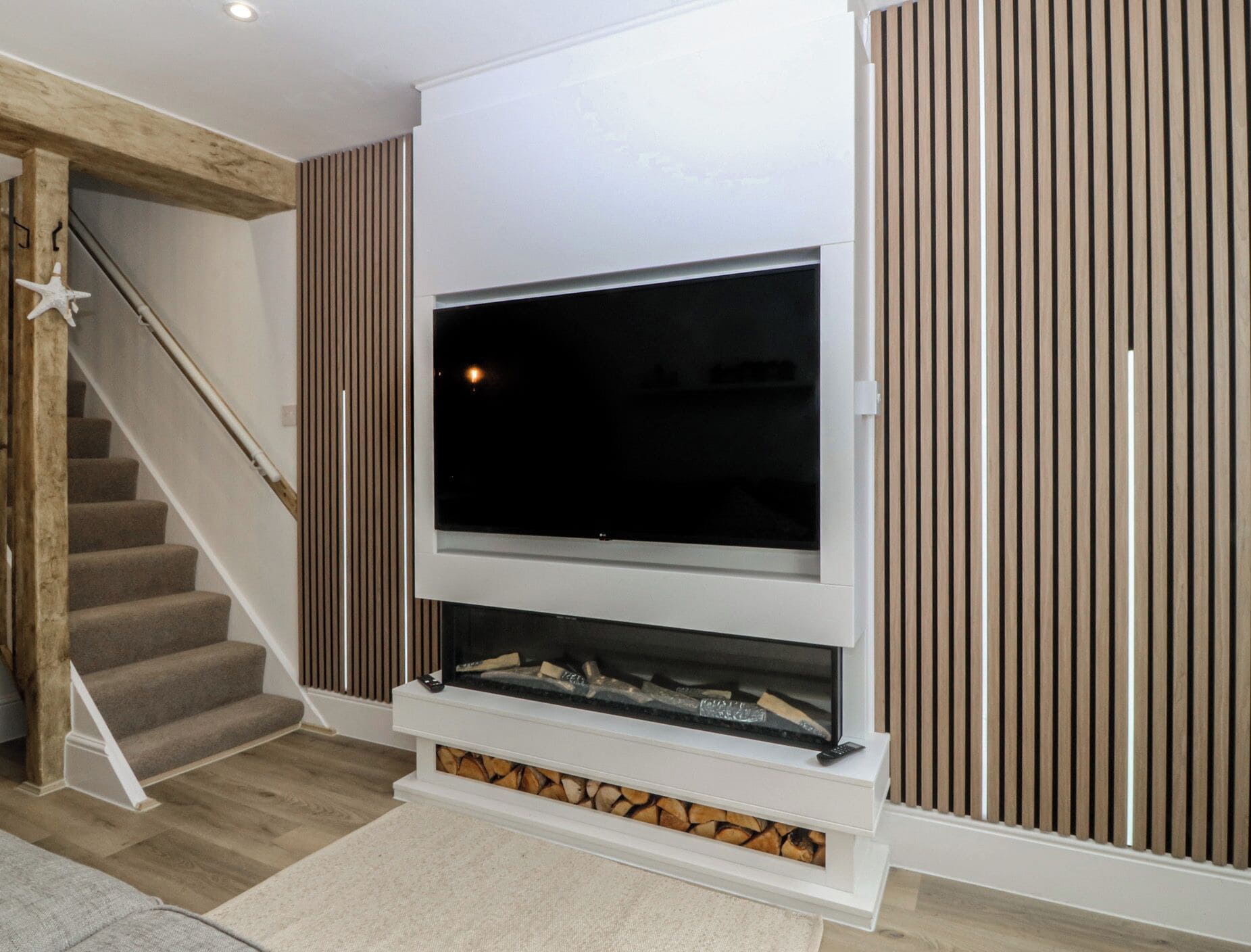 Ein modernes Wohnzimmer mit einem großen Flachbildfernseher an einer weißen Wand, einem Kamin mit Holzscheiten, Holzvertäfelung an den Wänden und einer Treppe mit einer Meerstern-Dekoration.