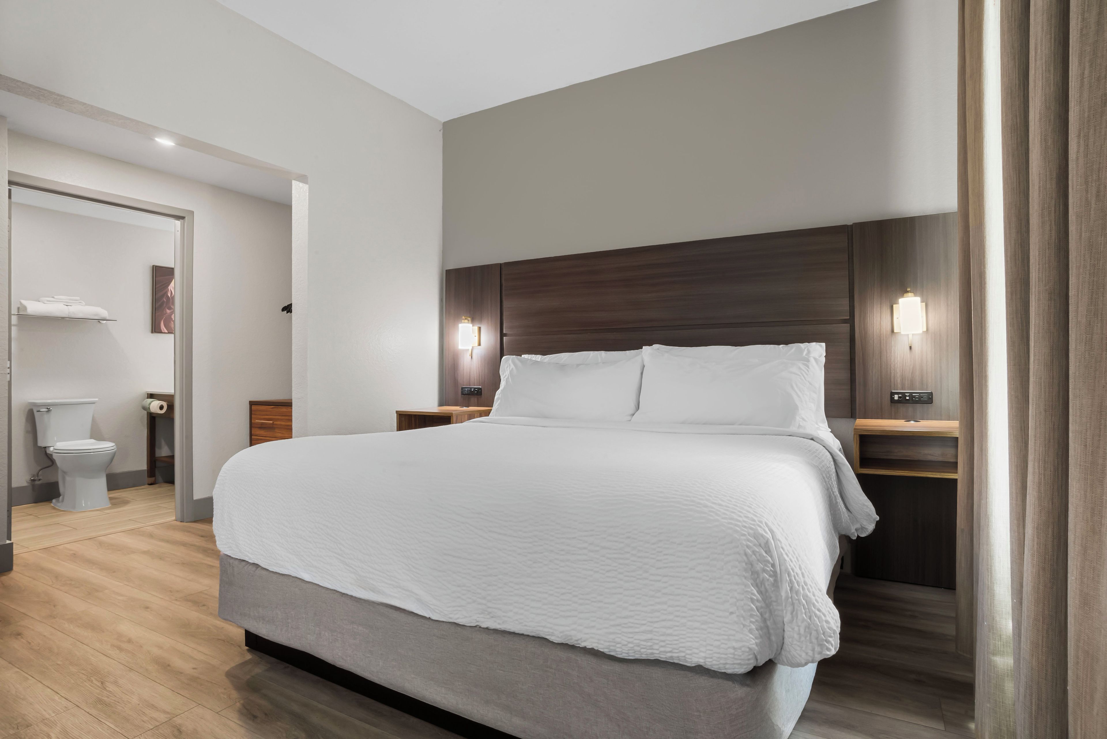 A modern hotel room with a large bed, wooden headboard, two bedside tables with lamps, a wooden floor, and a bathroom with a toilet and towel rack.