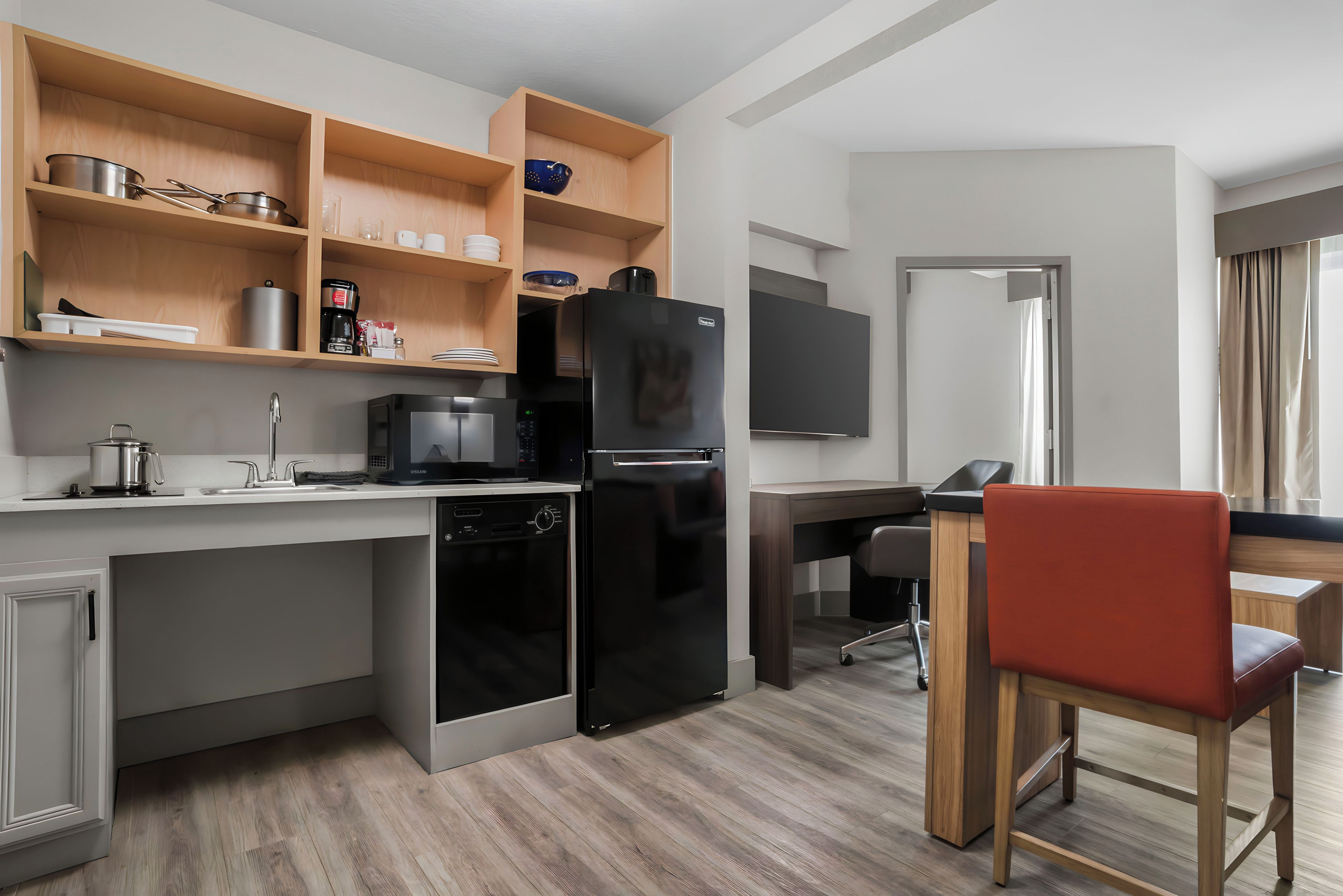 Cocina compacta con refrigerador, microondas y lavavajillas. Hay una mesa de madera con una silla, y un televisor de pantalla plana montado en la pared.