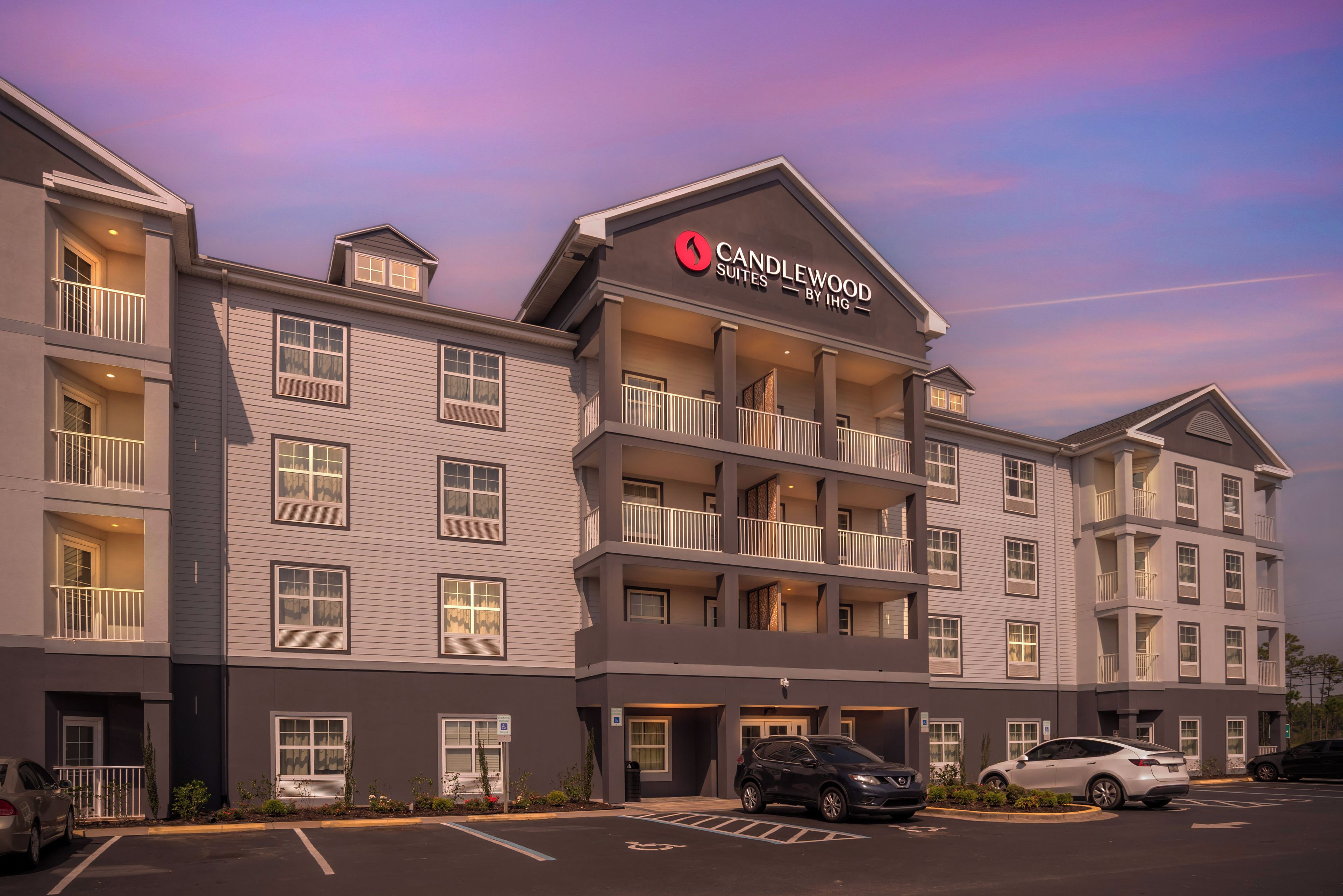 A multi-story hotel with a prominent 'Candlewood Suites by IHG' sign, multiple balconies, and a parking lot with several cars.