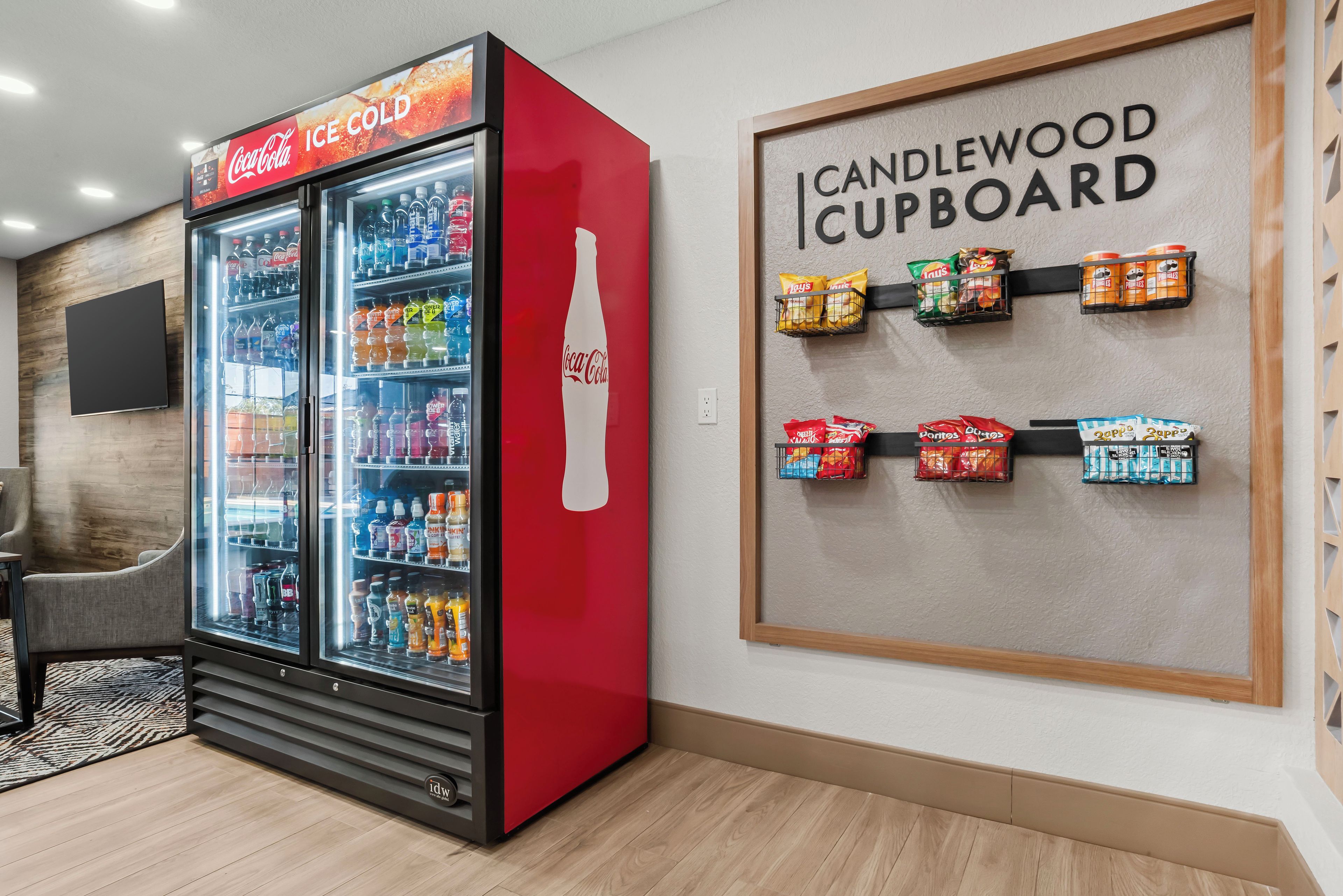 A room with a Coca-Cola refrigerator stocked with beverages, a wall-mounted sign reading 'CANDLEWOOD CUPBOARD,' and snack dispensers.