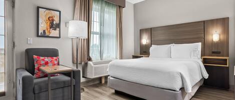 A modern hotel room with a bed, a chair, a desk, a window with curtains, and a painting on the wall.