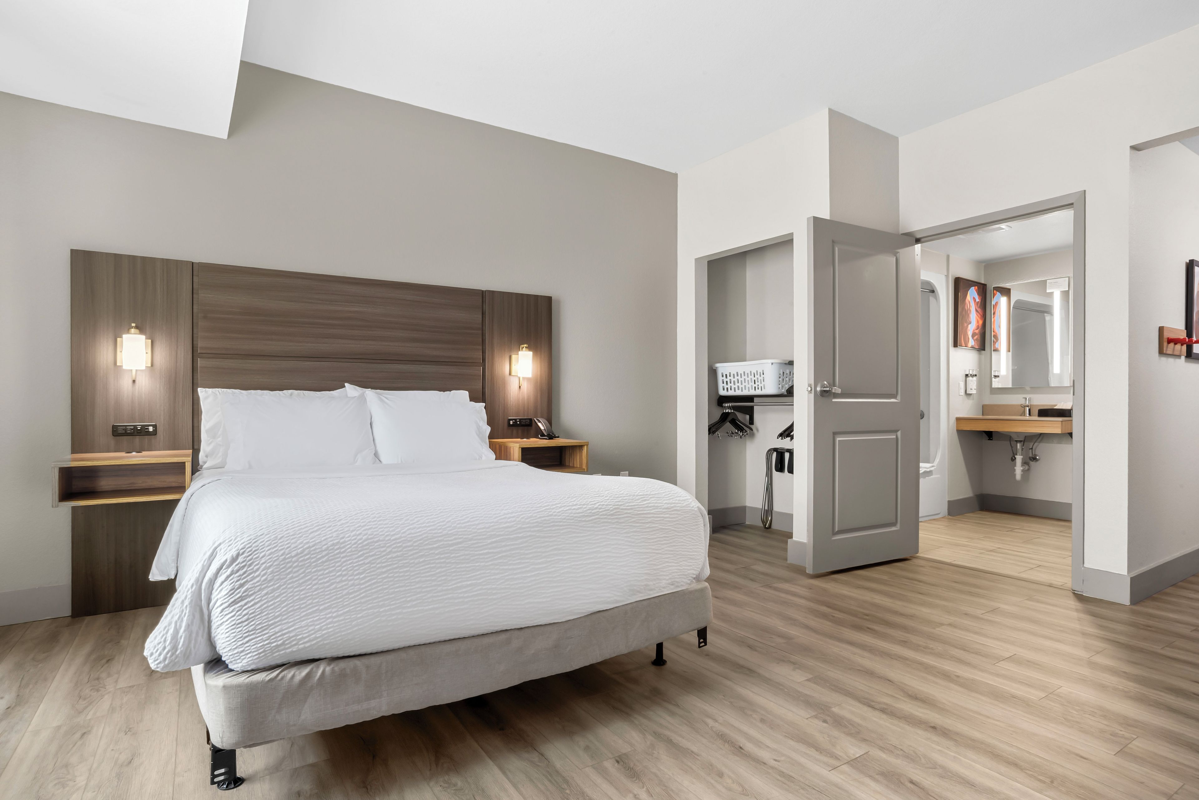 A modern hotel room with a large bed, bedside lamps, a wooden headboard, a small wooden nightstand, a bathroom with a sink and mirror, and a door leading to another room.