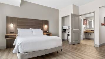 A modern hotel room with a large bed, bedside lamps, a wooden headboard, a small wooden nightstand, a bathroom with a sink and mirror, and a door leading to another room.