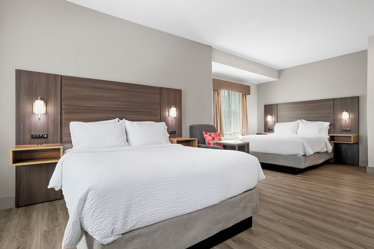 A hotel room with two beds, wooden headboards, bedside tables, and wall-mounted lighting.