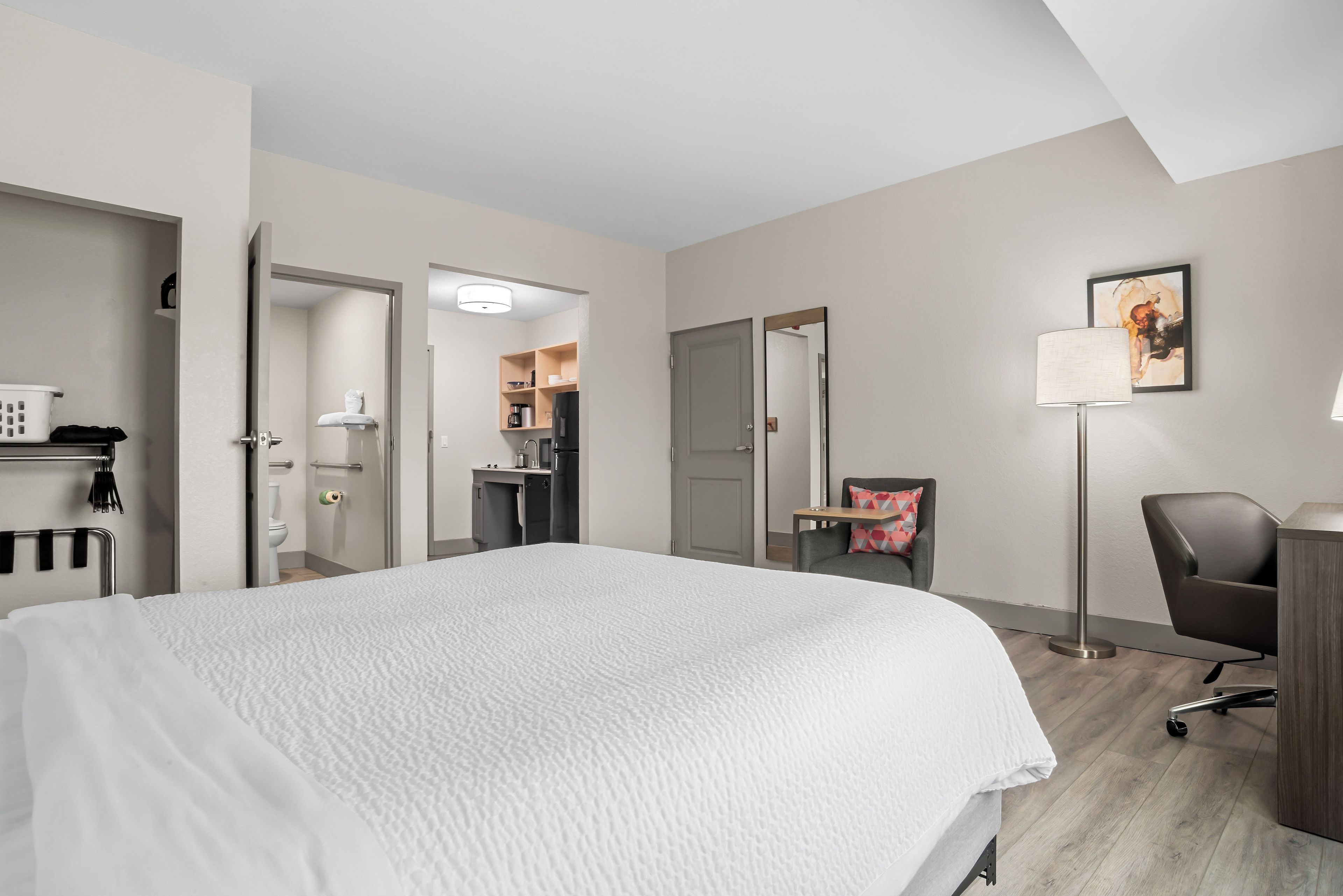 Quarto de hotel com cama, escrivaninha, poltrona, luminária de chão, um quadro e uma cozinha compacta ao fundo.