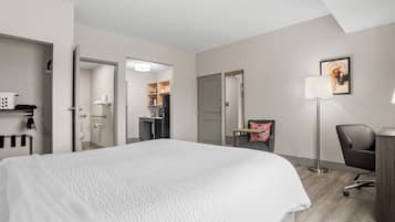Quarto de hotel com cama, escrivaninha, poltrona, luminária de chão, um quadro e uma cozinha compacta ao fundo.