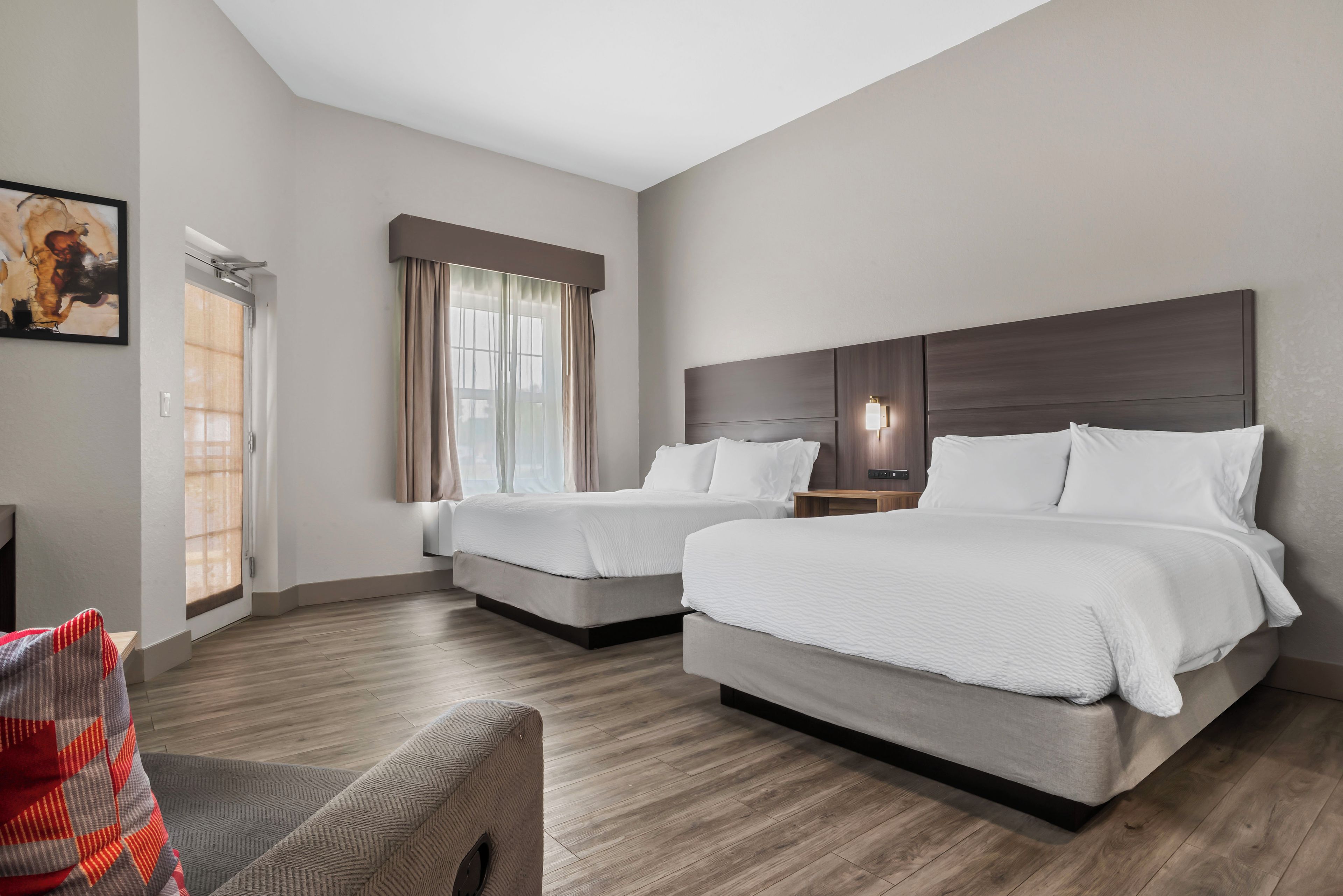 A hotel room with two beds, a large window with curtains, a painting on the wall, and a wooden floor.