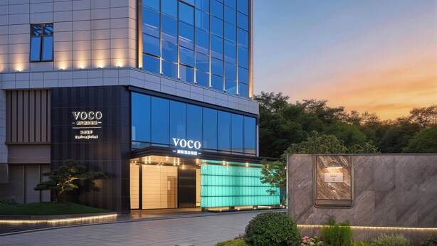 A modern building with a glass facade, a sign reading 'YOCO,' and a landscaped area with a small tree and bushes. - voco福州金汇 (Fuzhou)