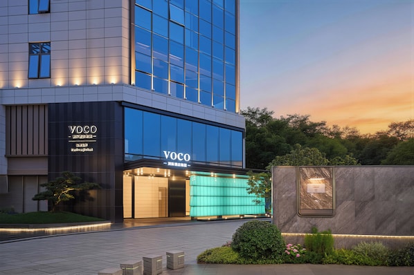 A modern building with a glass facade, a sign reading 'YOCO,' and a landscaped area with a small tree and bushes.
