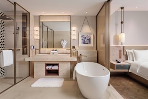 A modern bathroom with a large bathtub, a glass-enclosed shower, a vanity with a mirror, and a bedside table with a lamp.
