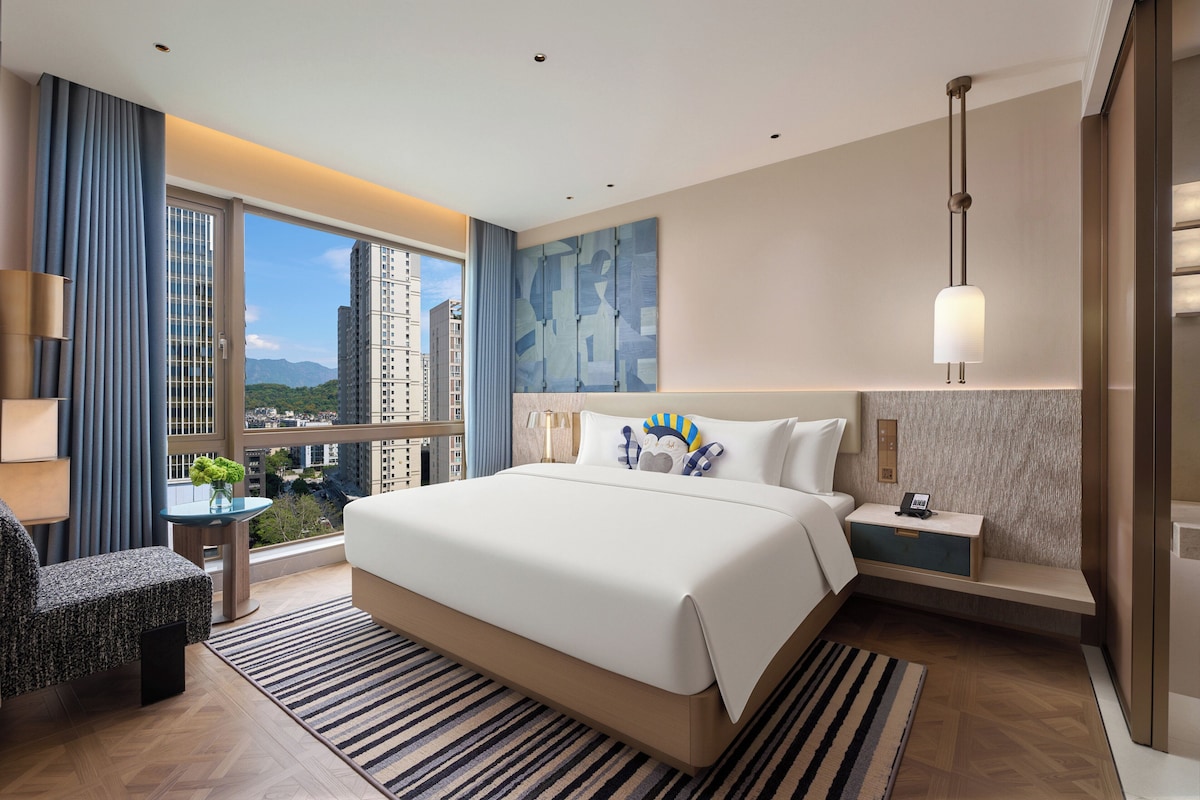 Una habitación de hotel moderna con una cama grande, un escritorio, una silla, vistas a la ciudad y un ventanal amplio.