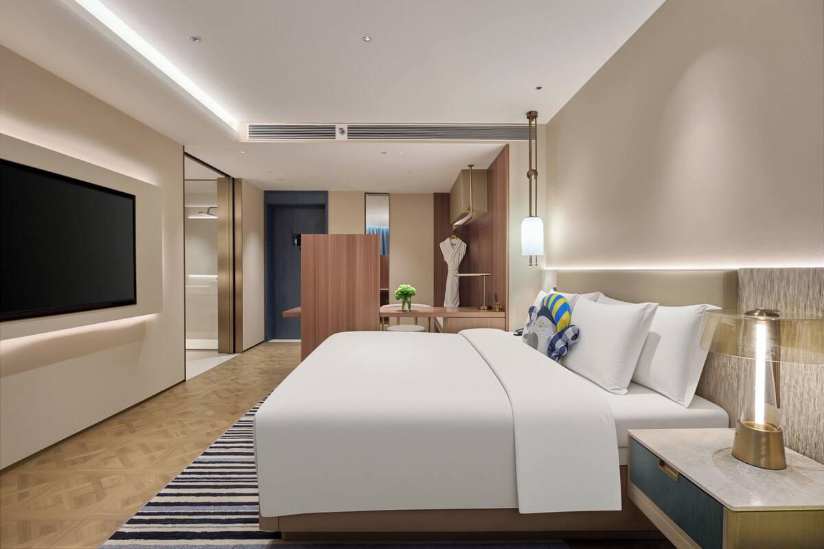 A modern hotel room with a large bed, bedside table, lamp, and a flat-screen TV.