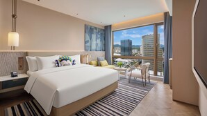 A modern hotel room with a large bed, a desk, a chair, a small table, and a view of the cityscape through large windows. - voco福州金汇 (Fuzhou)