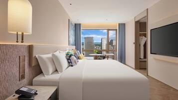 A modern hotel room with a large bed, a desk with a laptop, a TV, and a city view.