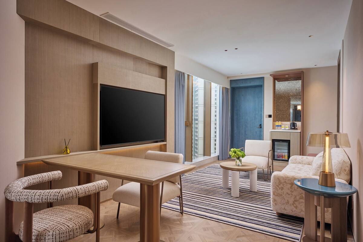A modern hotel room with a large flat-screen TV, a dining area with a table and chairs, a sofa, a small coffee table, and a lamp.